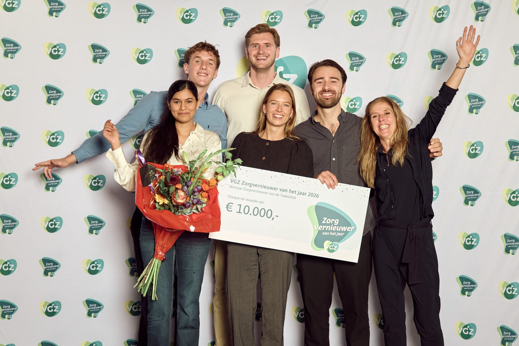 Het PAVA-team voor de fotowand van VGZ met de cheque van 10.000 euro voor Winnaar Zorgvernieuwer van de Toekomst, met bloemen en een arm in de lucht